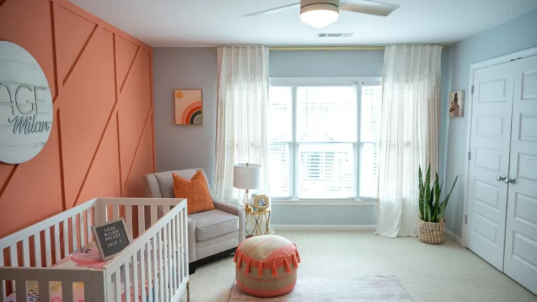 Boho Nursery with Peach Fuzz accent wall. Light Blue gray on other walls. Peach Fuzz accents. White sheer curtains, white blinds, white trim, white crib, white carpet, large snake plant in seagrass basket in the corner