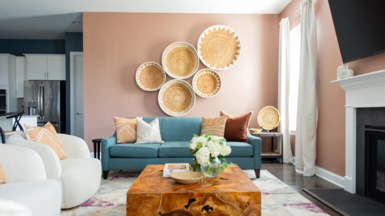 Boho Styled Living Room featuring a Peach Fuzz accent wall and a basket wall of 5 different size flat baskets. Teal couch. White chairs. Both having Peach Fuzz color accent pillows. Live Edge wood coffee table in center. White curtains & white trim around bottom of walls and fireplace. Fireplace and TV are black. Dark floors with a white rug accented in magenta and peach tones.