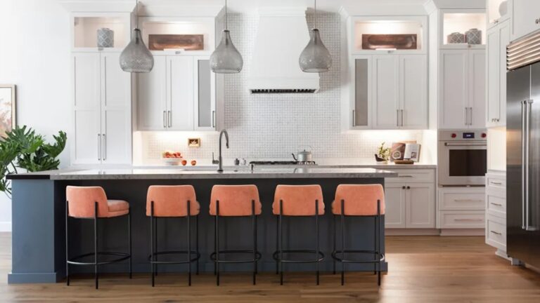 White modern kitchen with a large island in the middle. Bottom of island is painted a dark muted blue/gray. Peach Fuzz color accent barstools around island.