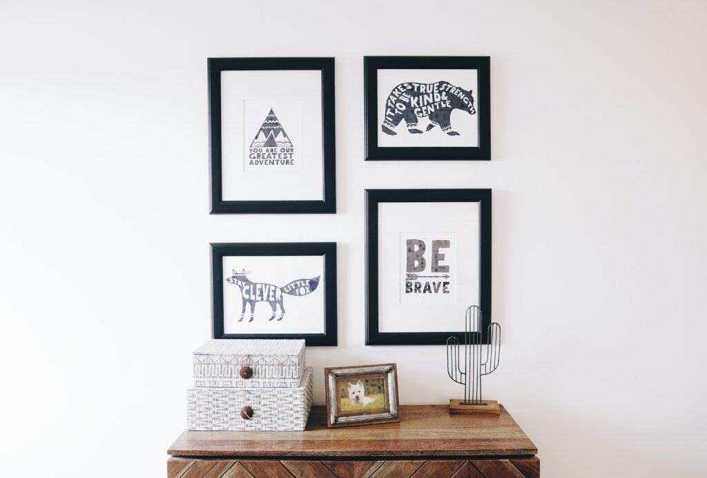 Black and white framed pictures hanging on a wall above a wood desk with a cactus art piece, a box, and picture that is framed.