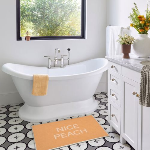 White bathroom with black and white tiled patterned floor. White tub, Plants with Peach Fuzz accents. Bath Mat that has text that says "Nice Peach" color of bathmat is Peach Fuzz
